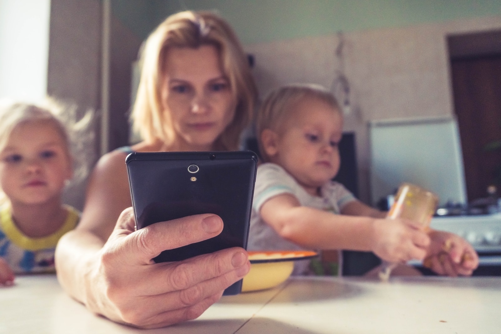 busy woman in white shirt holding black ipad caring for two young children